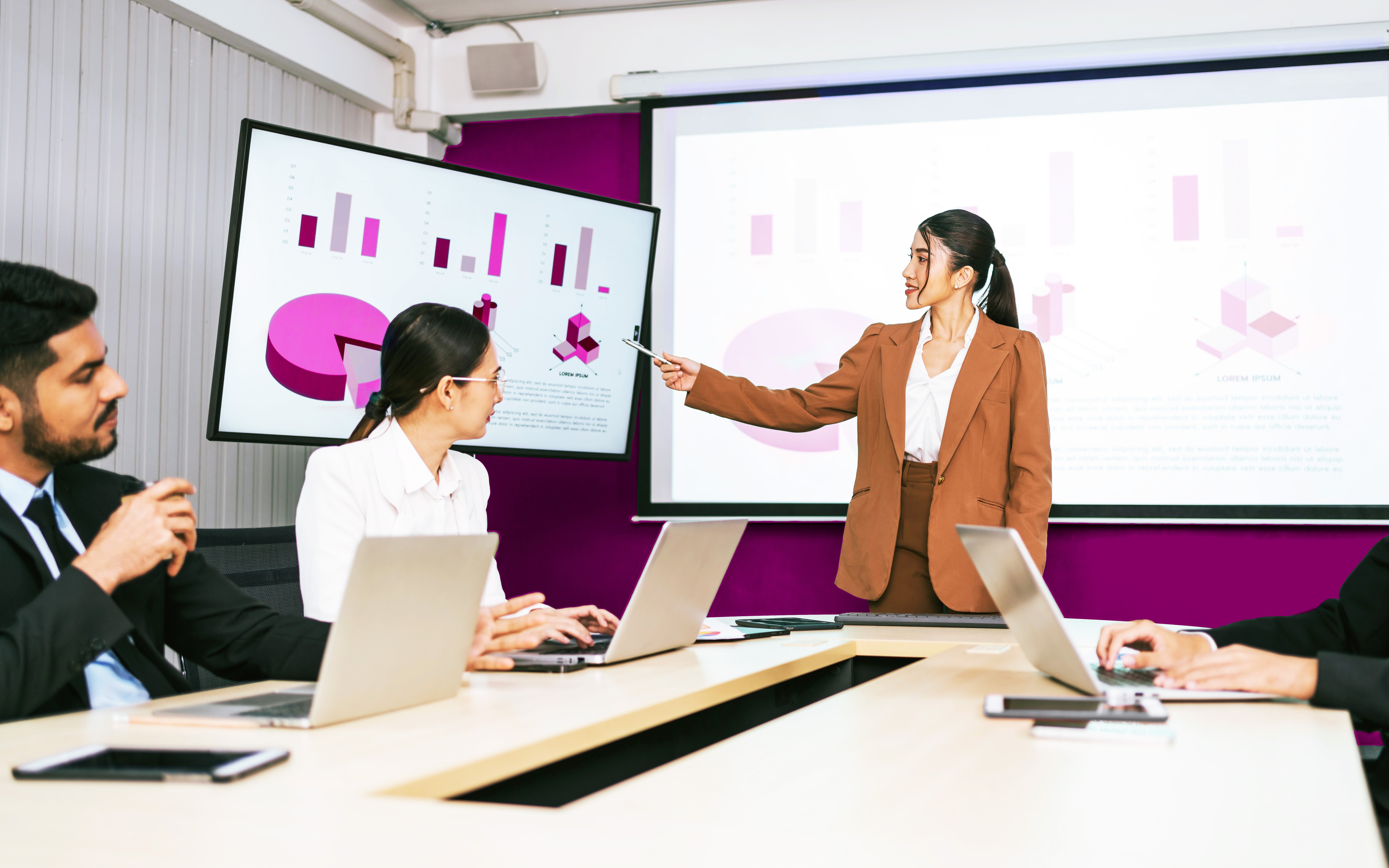 A cheerful and confident Asian businesswoman stands, present bar charts data from projector screen to her office colleagues